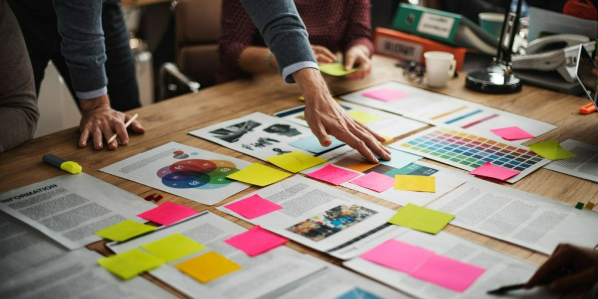 Imagem de um grupo de pessoas colaborando em um projeto, com várias mãos apontando e organizando papéis, gráficos, notas adesivas coloridas e uma paleta de cores espalhadas sobre uma mesa de madeira. A cena sugere um ambiente de brainstorming ou planejamento criativo.