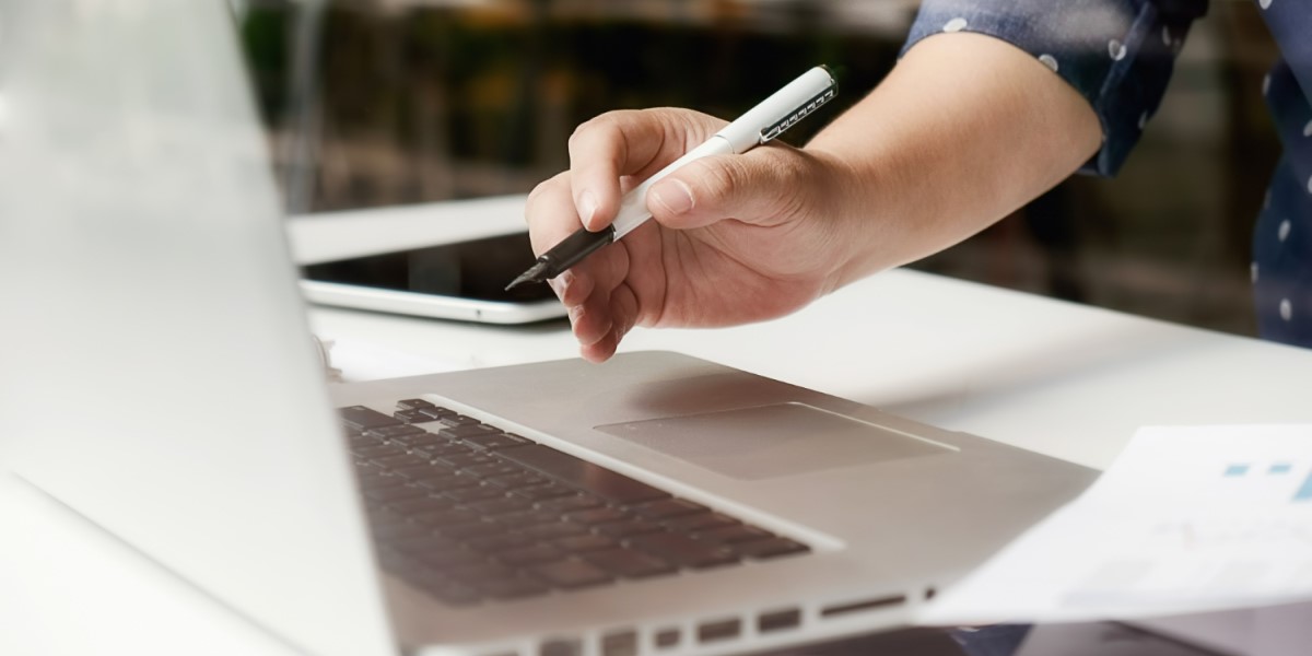 Pessoa escrevendo com uma caneta em um bloco de notas ao lado de um laptop aberto.