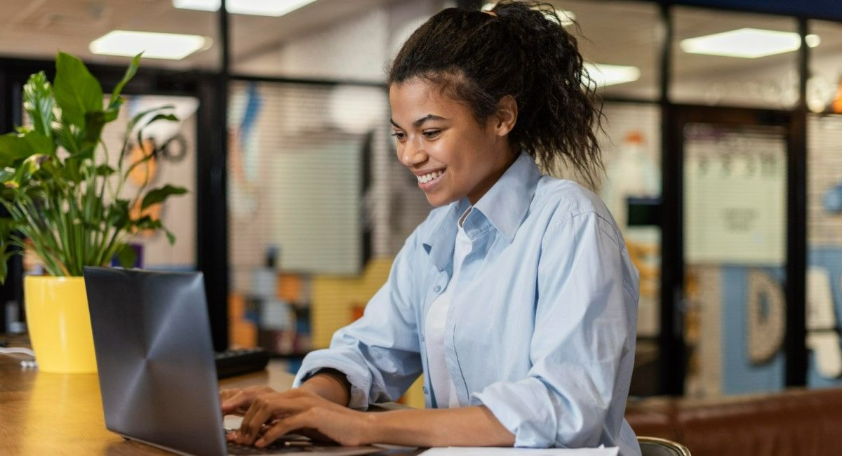 Mulher sorridente trabalhando em um laptop em um escritório moderno, com uma planta ao lado e ambiente de trabalho ao fundo.