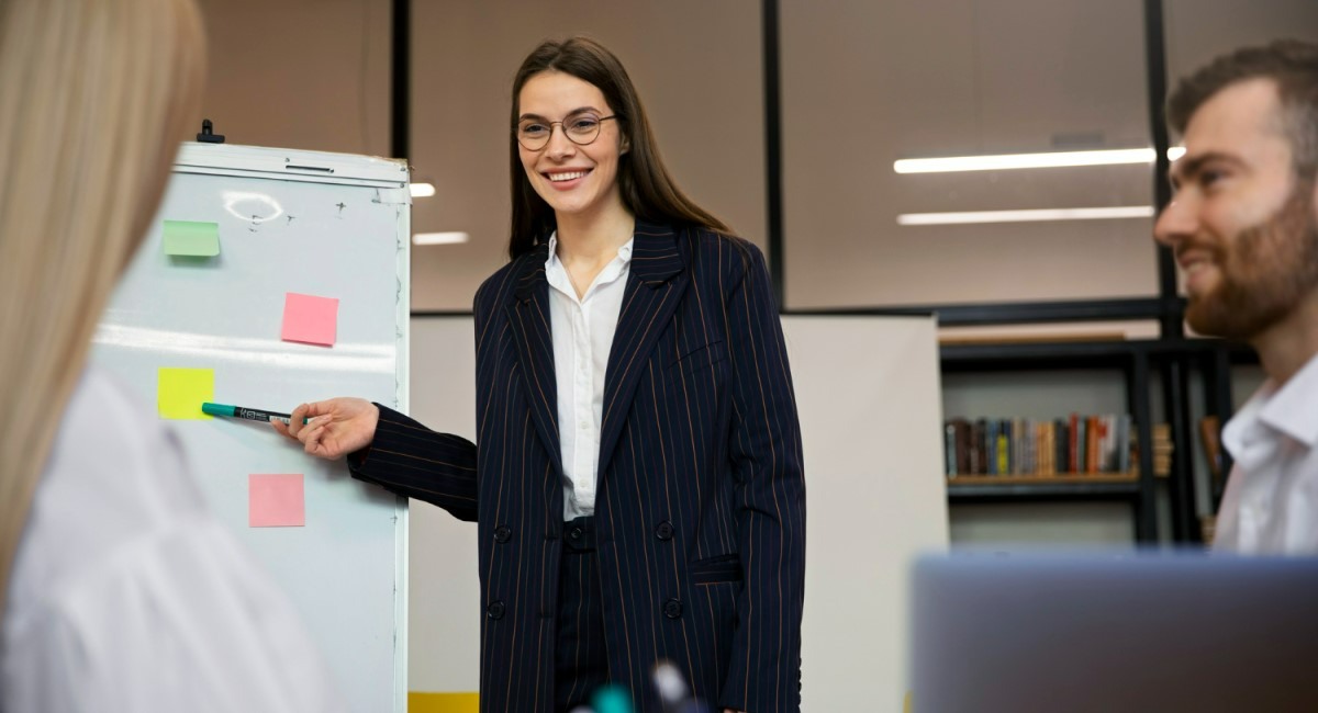 Mulher sorridente em uma apresentação no escritório, usando blazer listrado, apontando para um quadro branco com post-its coloridos, enquanto colegas assistem atentamente.