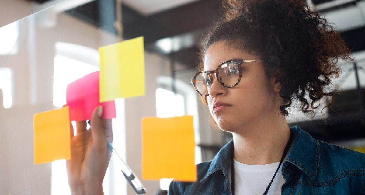 Mulher jovem olhando atentamente para post-its coloridos em um vidro, em um ambiente de escritório moderno.