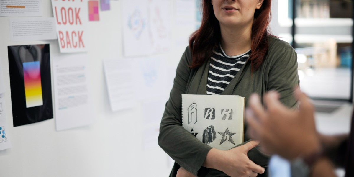 Mulher segurando caderno com desenhos de ícones, em pé em frente a uma parede de inspirações com papéis e cartazes, em um ambiente de escritório colaborativo.