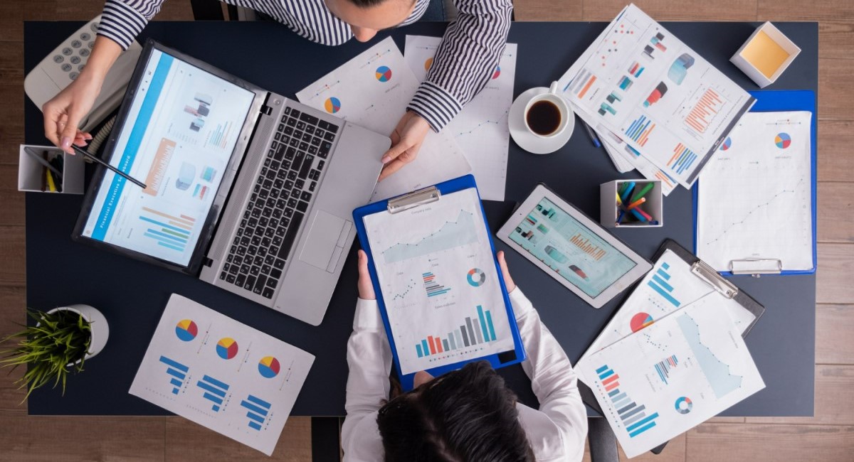 Vista de cima de uma mesa de escritório com gráficos, planilhas e laptops, onde duas pessoas trabalham analisando dados e discutindo relatórios