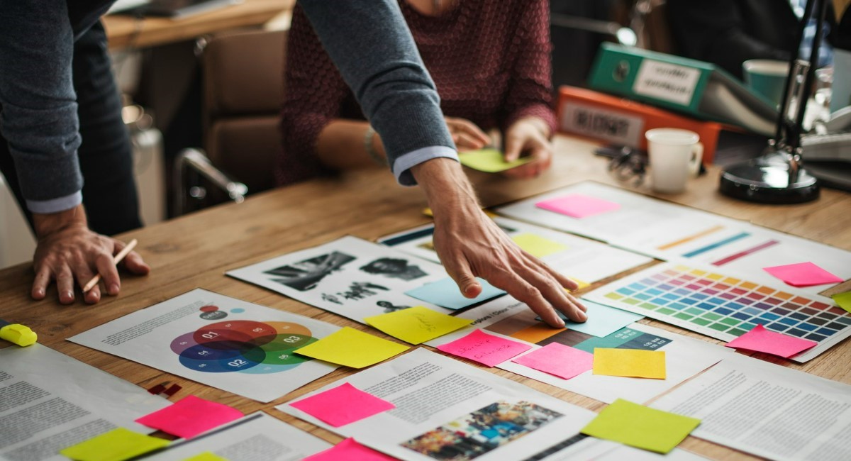 Pessoas organizando documentos, fotos e amostras de cores em uma mesa coberta de papéis e post-its coloridos durante uma sessão de brainstorming ou planejamento criativo.