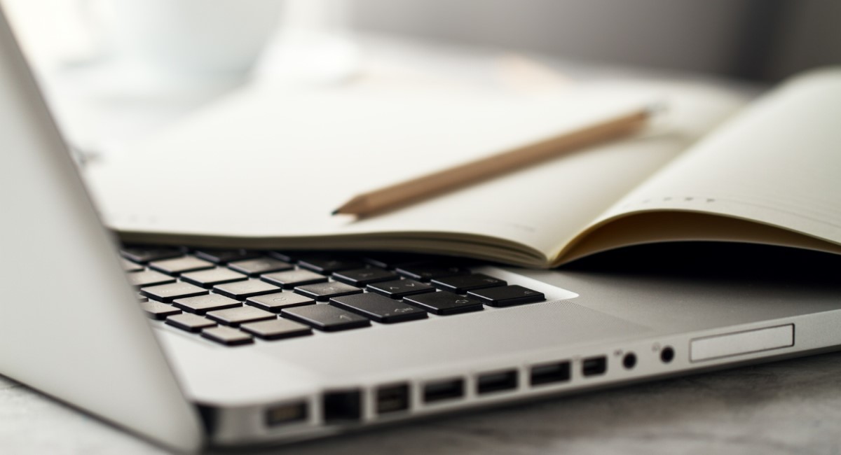 Close-up de um teclado de laptop com um caderno aberto e um lápis em cima, sugerindo um espaço de trabalho para escrita ou estudo