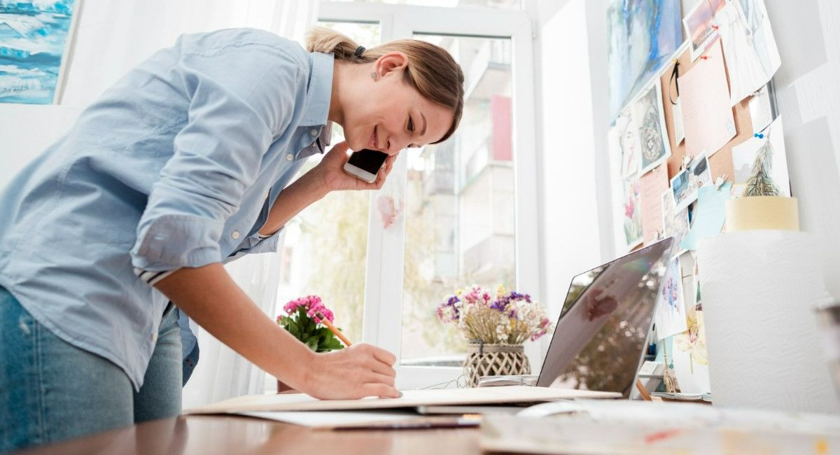 Mulher trabalhando em um escritório, falando ao telefone enquanto faz anotações em um caderno. Ela está em pé diante de uma mesa com um laptop e papéis, em um ambiente iluminado e decorado, representando multitarefa, produtividade e trabalho remoto.