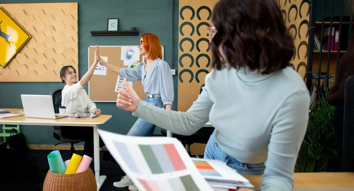 Três mulheres colaborando em um ambiente de escritório moderno. Uma delas segura uma paleta de cores, enquanto as outras duas discutem ideias próximas a um quadro de avisos. O cenário representa brainstorming, criatividade e trabalho em equipe em um ambiente descontraído e inspirador.