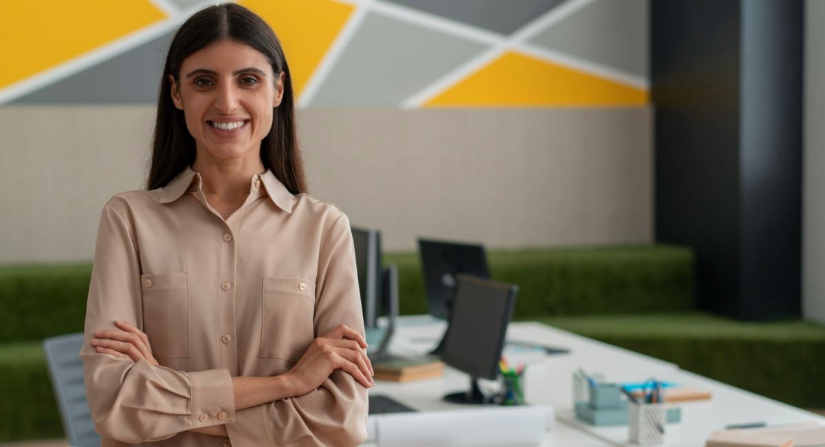 Mulher jovem, de cabelos longos e escuros, sorrindo e de braços cruzados em um ambiente de escritório moderno, com computadores e decoração em tons de amarelo, cinza e verde ao fundo.