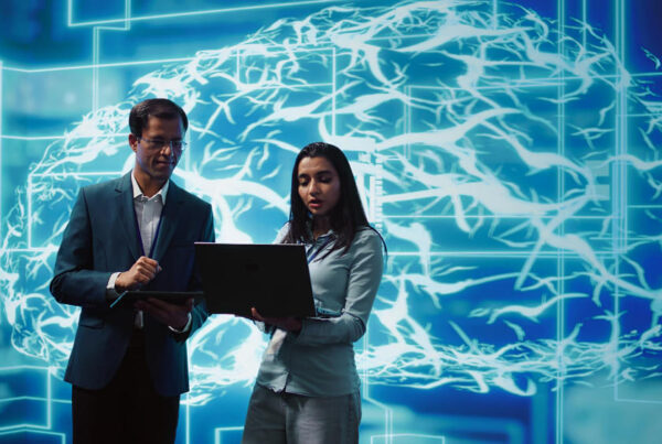 Dois profissionais analisam informações em um notebook e um tablet, em frente a uma representação digital de um cérebro com circuitos tecnológicos, simbolizando o uso da inteligência artificial e da análise de dados no ambiente corporativo.