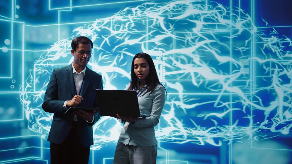 Dois profissionais analisam informações em um notebook e um tablet, em frente a uma representação digital de um cérebro com circuitos tecnológicos, simbolizando o uso da inteligência artificial e da análise de dados no ambiente corporativo.